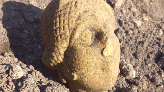 A stone Roman head carving lying on a pebbled ground. It is pale yellow and has its eyes closed and a damaged nose.