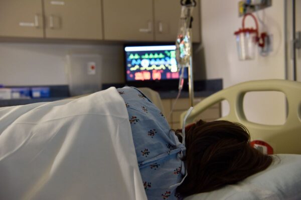 A patient with dark hair wearing a blue hospital gown lies in a hospital bed. There is various hospital equipment and a monitor in the frame.