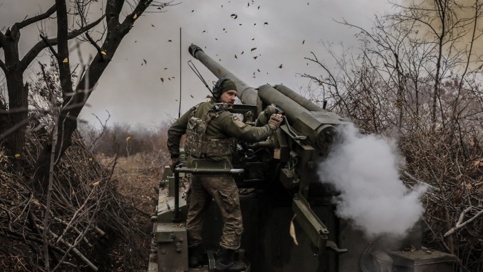 A Ukrainian serviceman operates a 2S5 152 mm self-propelled howitzer, firing towards Russian positions. The scene is set in a wooded area, with smoke and leaves visible around the artillery.