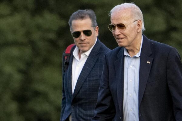 Two White men wearing sunglasses and navy suits are walking in a field.