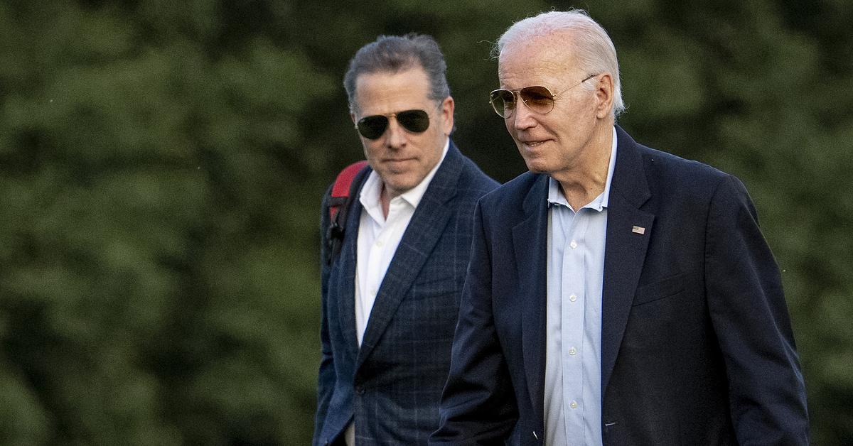 Two White men wearing sunglasses and navy suits are walking in a field.