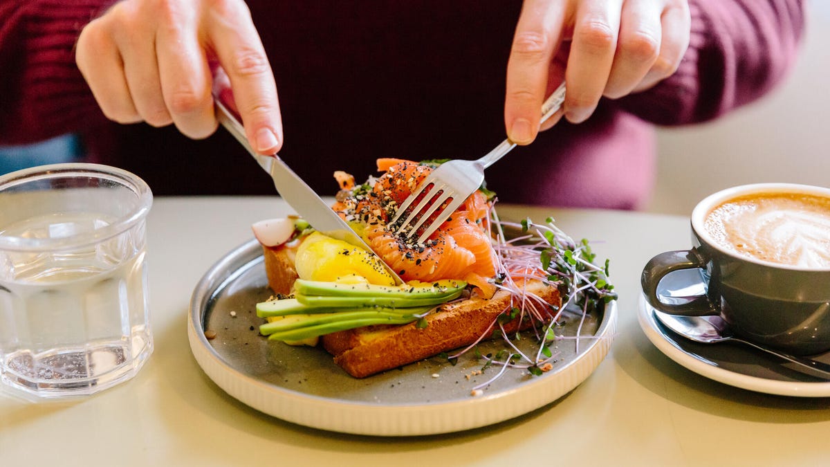 A man&apos;s hands cutting a salmon and avocado flatbread with a knife and fork.