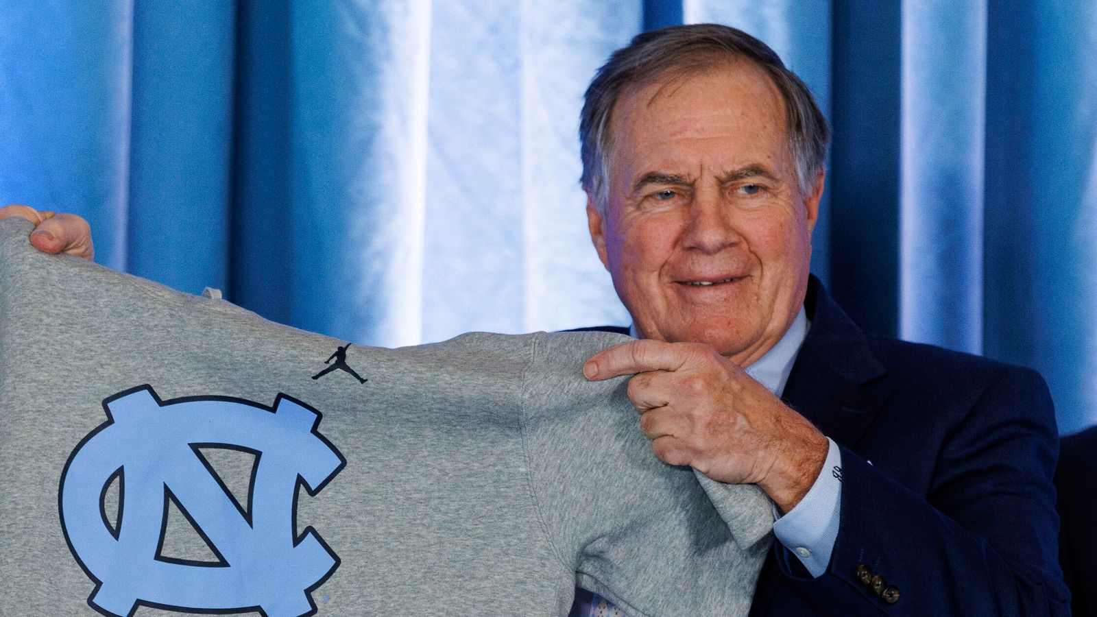 New North Carolina football coach Bill Belichick holds up a UNC branded sleeveless hoodie presented to him during an NCAA college football press conference announcing his hiring, Thursday, Dec. 12, 2024, in Chapel Hill, N.C. (AP Photo/Ben McKeown)