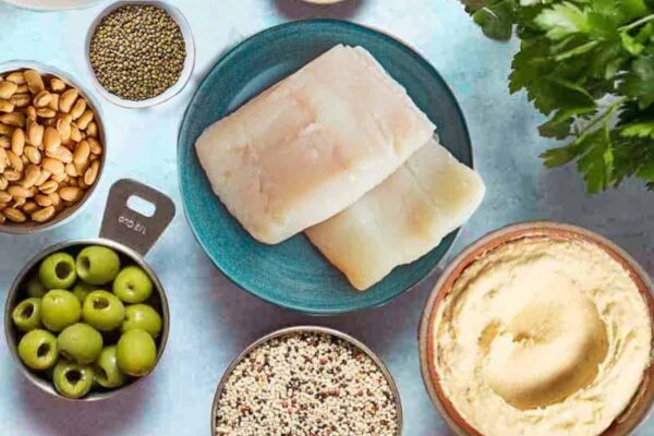 An overhead photo of several Mediterranean diet friend foods including eggs, fish, clams, mussels, feta cheese, aleppo pepper, lentils, quinoa, beans, olives, tomatoes, lemons, cucumber, hummus and parsley.