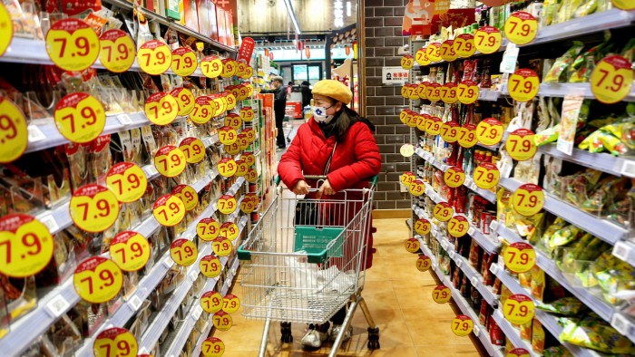 A customer wearing a face mask and a red coat browses through supermarket aisles filled with products marked with bright yellow and red sale signs in Lianyungang, Jiangsu province, China. She pushes an empty shopping cart