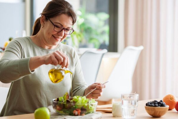 A woman with brown hair in a ponytail and glasses pouring olive oil onto a fresh salad.