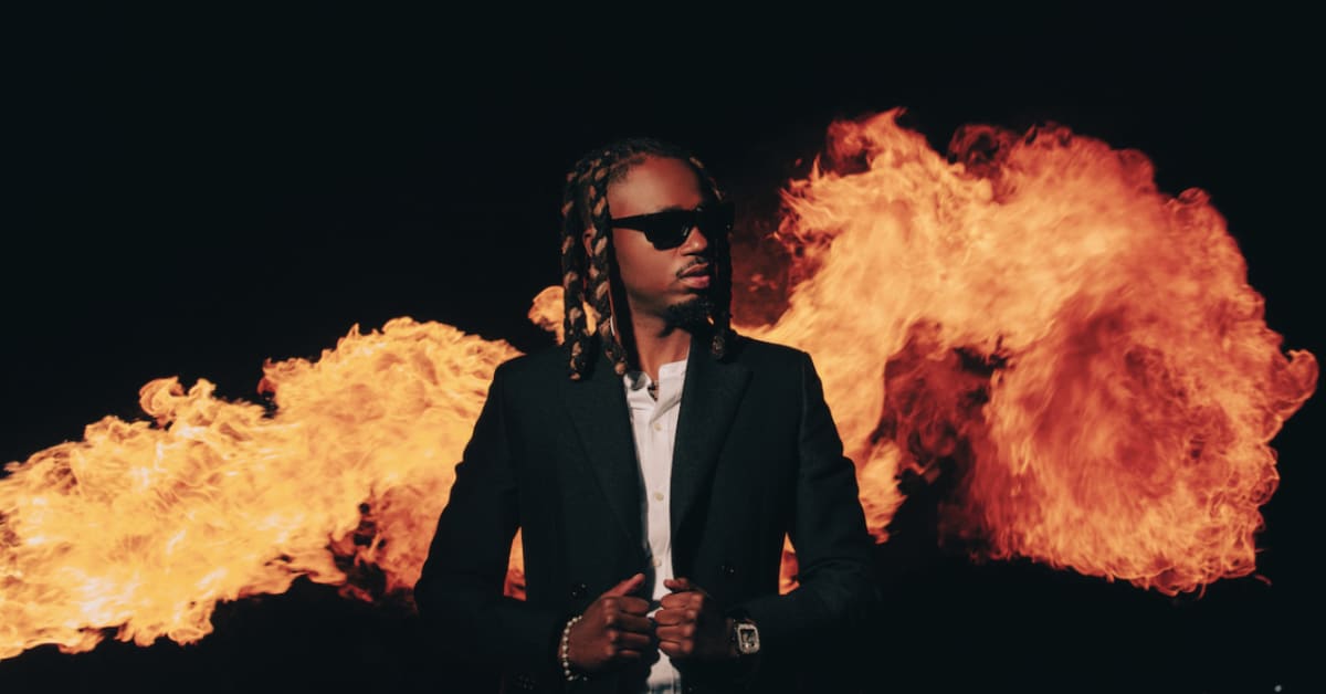 Metro Boomin with braided hair and sunglasses in front of large flames, wearing a dark suit and white shirt, looking to the side.