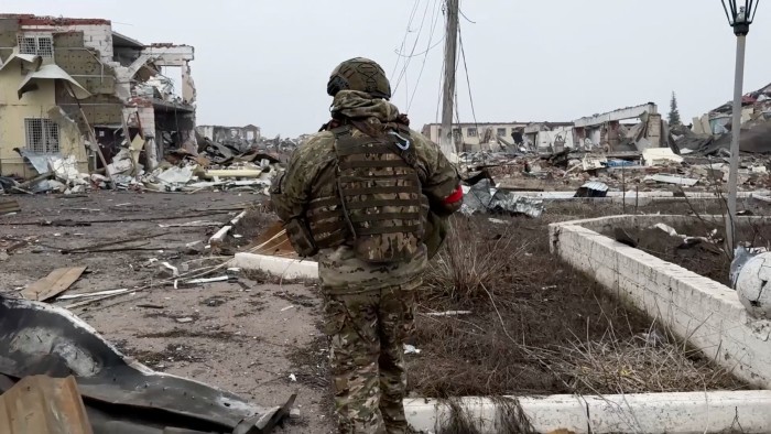 A Russian soldier, wearing camouflage and tactical gear, walks along a debris-strewn street