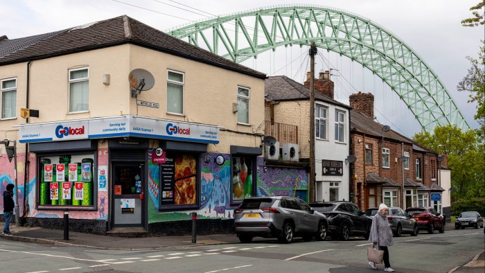 A woman walks across Waterloo Road in Runcorn, England. The street features a corner shop with colourful murals, parked cars, and the arch of a large bridge in the background.