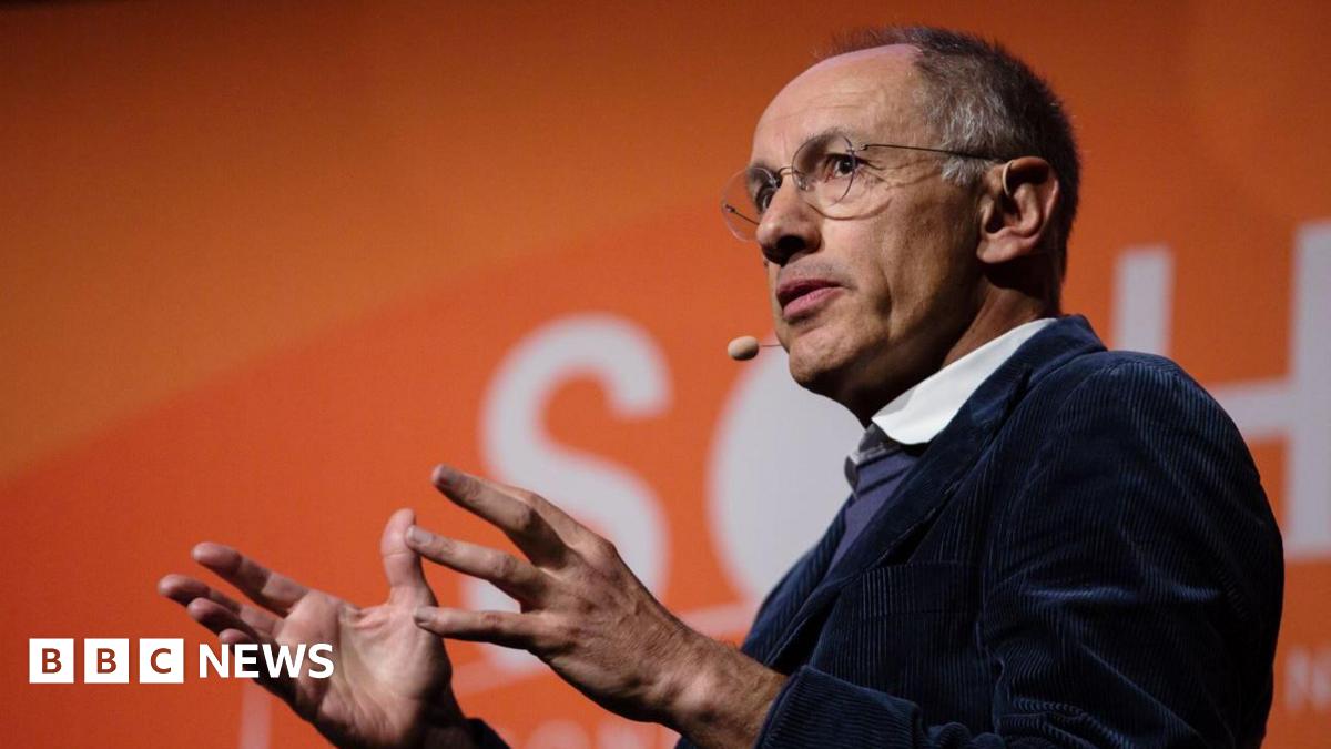 Michael Moritz is on stage, speaking with his hands in front of him with an orange background behind him. He has a microphone headset by his mouth and is wearing a navy corduroy jacket with a blue jumper and white shirt underneath.