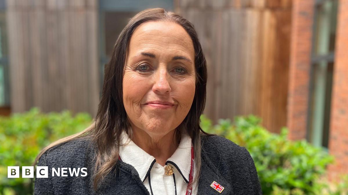 Councillor Joanne Monk photographed outside the council buildings. She has long brown hair and is wearing a light shirt, dark jacket and a lanyard. She has a Union Jack pin badge on her jacket lapel and is smiling at the camera. There is a low green bush behind her.