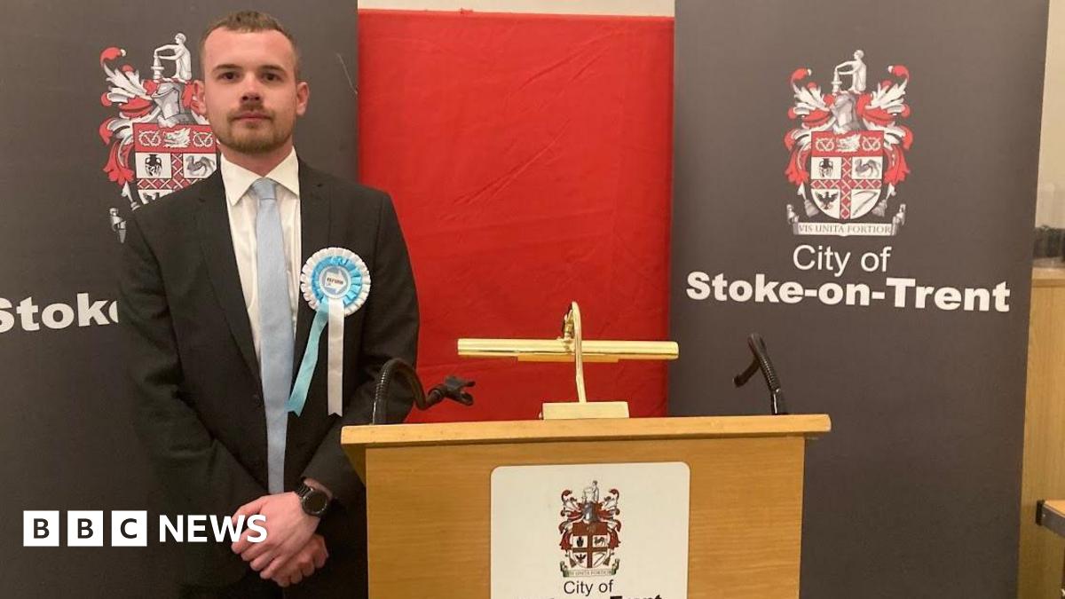 A man stands next to a podium at a by-election count. He has his hands clasped in front of him and he's wearing a blue and white Reform rosette.