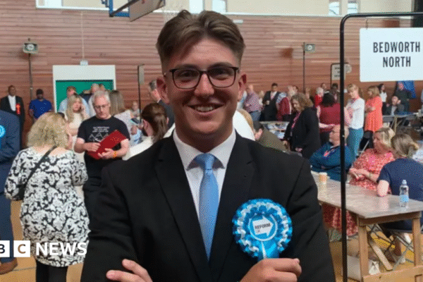 George Finch pictured smiling while crossing his arms. He is wearing wide round black framed glasses and is in a black suit jacket with a white shirt underneath and a blue tie.