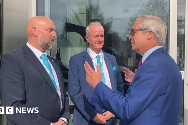 Eddie McDonald with very short hair and grey beard, Martin Griffiths with short white hair and Nigel Farage with short white hair. They are all wearing blue jackets and light blue ties. Farage has both hands up as he is explaining something. There are glass doors behind them.