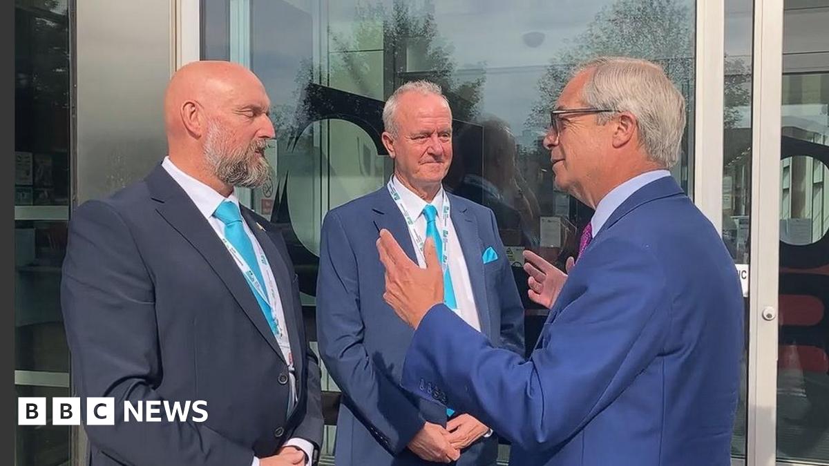 Eddie McDonald with very short hair and grey beard, Martin Griffiths with short white hair and Nigel Farage with short white hair. They are all wearing blue jackets and light blue ties. Farage has both hands up as he is explaining something. There are glass doors behind them.
