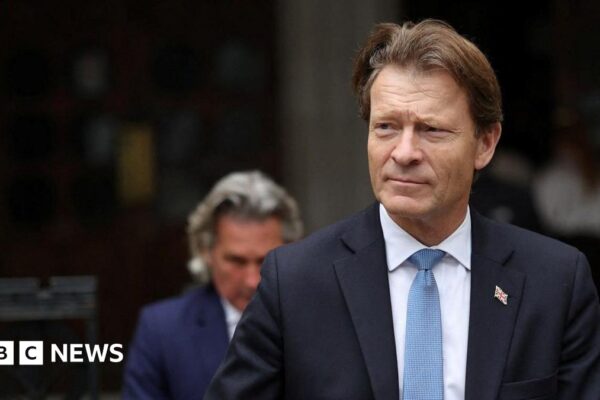 A man dressed smartly in a navy suit, white shirt and blue tie walks down a street in front of a dark building. He has brown, swept-back hair and wears a Union flag lapel badge. A second man, who is out of focus, follows.