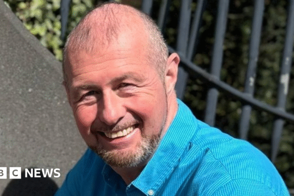 Justin Bellhouse is wearing a light blue, buttoned up shirt. He has thinning hair and a goatee beard. He is smiling.