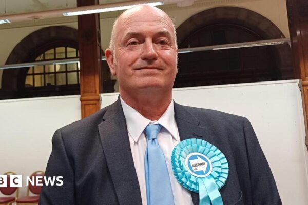 A middle-aged man wearing a light blue rosette and a suit stands in a public hall of some kind. His tie is also light blue.