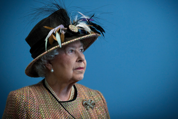 Queen Elizabeth wearing a feathered hat and tan jacket standing in front of a blue wall