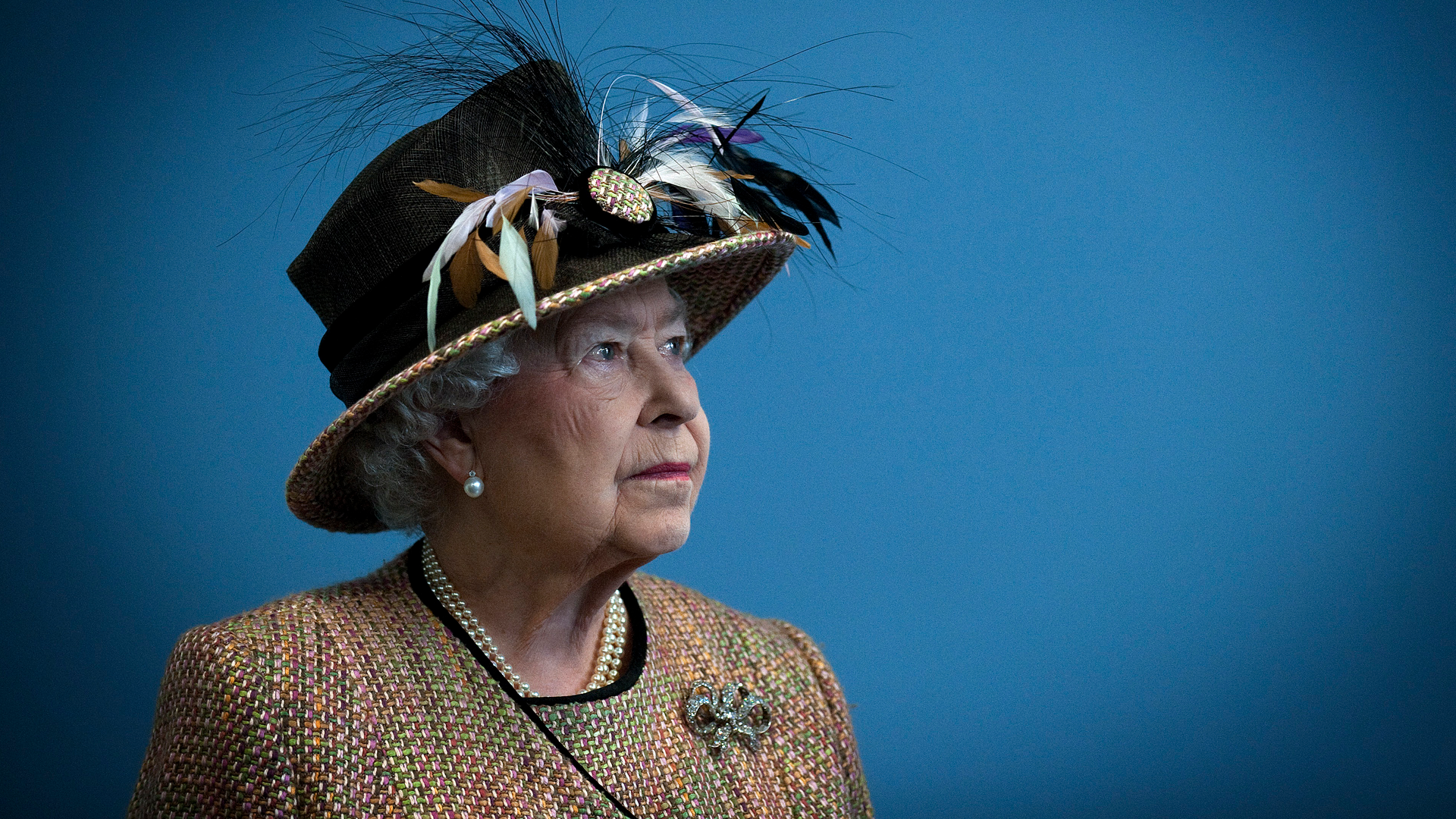 Queen Elizabeth wearing a feathered hat and tan jacket standing in front of a blue wall