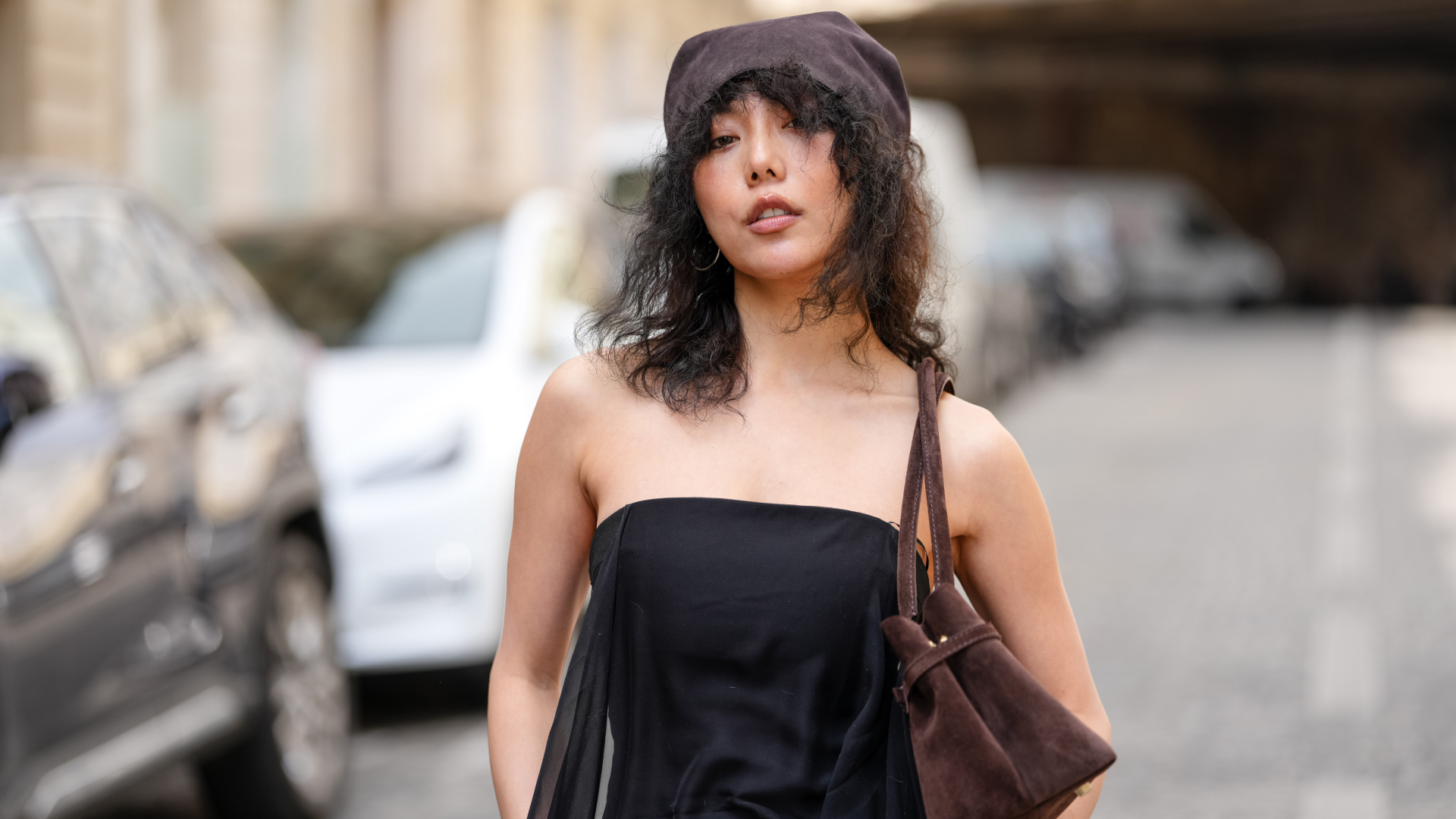 Xiayan Guo wears a brown suede beret with medium-length curly black hair and a middle part, styled with a strapless black jumpsuit featuring a sheer draped overlay. A dark brown suede shoulder bag and silver bangles complete the visible accessories. This is a mid shot, during a street style fashion photo session, on May 07, 2025 in Paris, France. (Photo by Edward Berthelot/Getty Images)