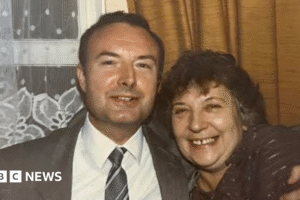 An old photograph of Mr Hill with a woman. Both are smiling at the camera. Mr Hill has short dark hair and is wearing a grey suit and a grey tie, while the woman is in a dark blouse.