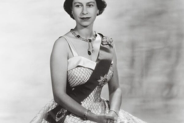 The Largest Exhibition of Queen Elizabeth II’s Fashion to Be Displayed at Buckingham Palace A formal portrait of a woman wearing a crown and sash with a jeweled necklace and elegant gown against a neutral background.