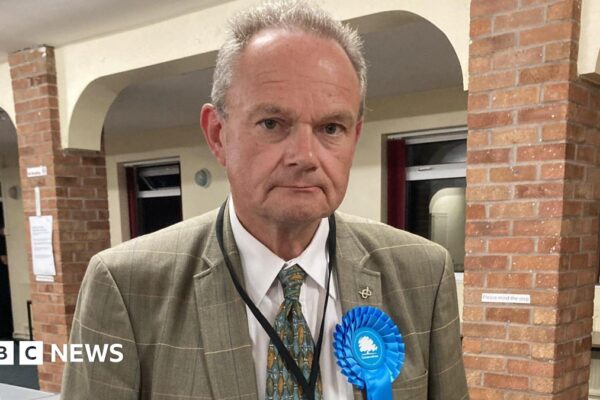 A man with short grey hair, a grey jacket, green tie and a blue Conservative Party rosette, stands in a hall with brick pillars and a white wall with windows behind him.