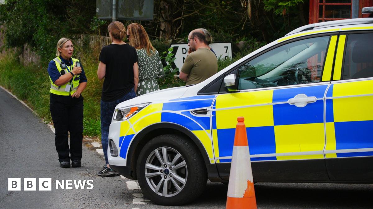 A female police officer in uniform talks to local residents standing next to a police car in Wheddon Cross in Somerset after a coach crash