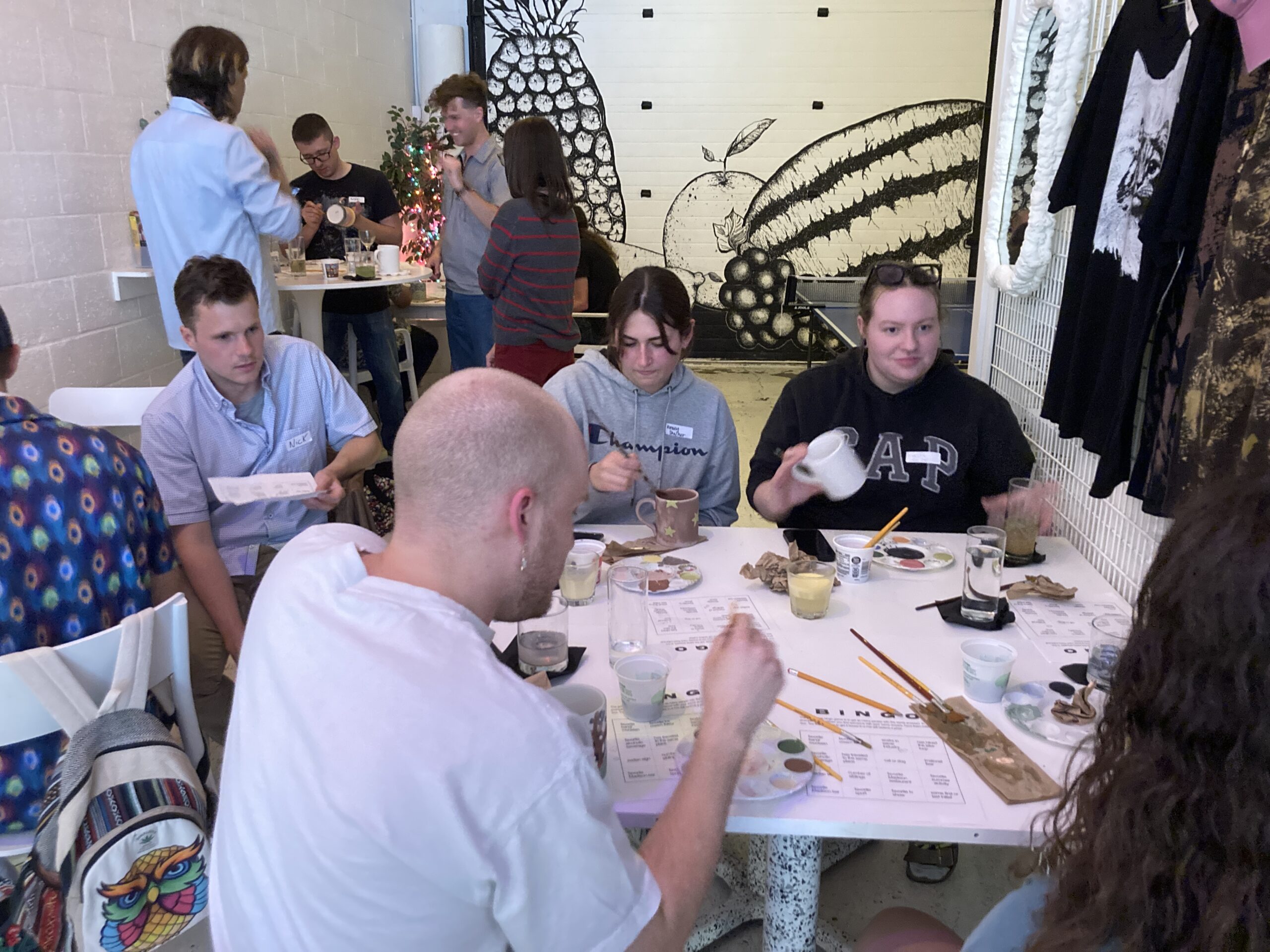 People sit around a table engaged in an art activity, using brushes and paints. Other people stand and talk in the background. Black and white fruit murals are on the wall.
