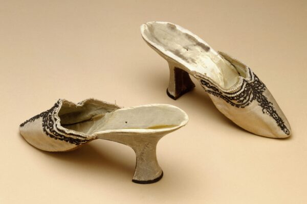 Pair of vintage heeled shoes with intricate black lace embellishments on a beige background.