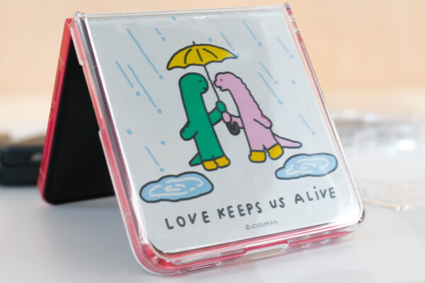 Samsung Galaxy Z Flip 7 in coralred in a cute case that says Love Keeps Us Alive and shows two dinosaurs sharing an umbrella
