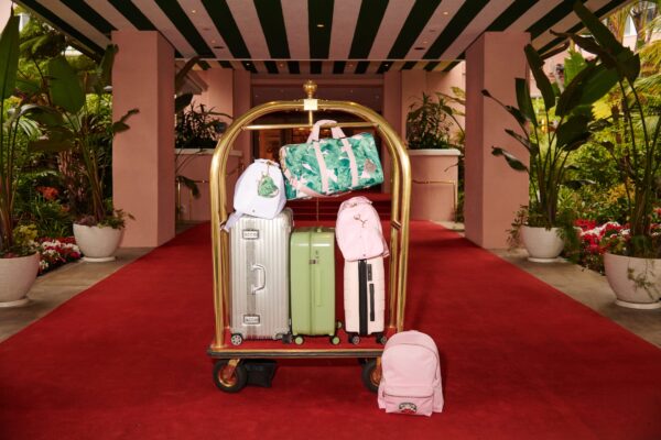 These Hotel-Fashion Exclusives Make Summer’s Chicest Collectibles Luggage cart with assorted bags and suitcases standing on a red carpet in front of a hotel entrance with striped awning.