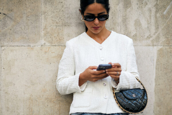 woman wearing white tweed jacket, sunglasses, and jeans looking at her phone and carrying a chanel bag