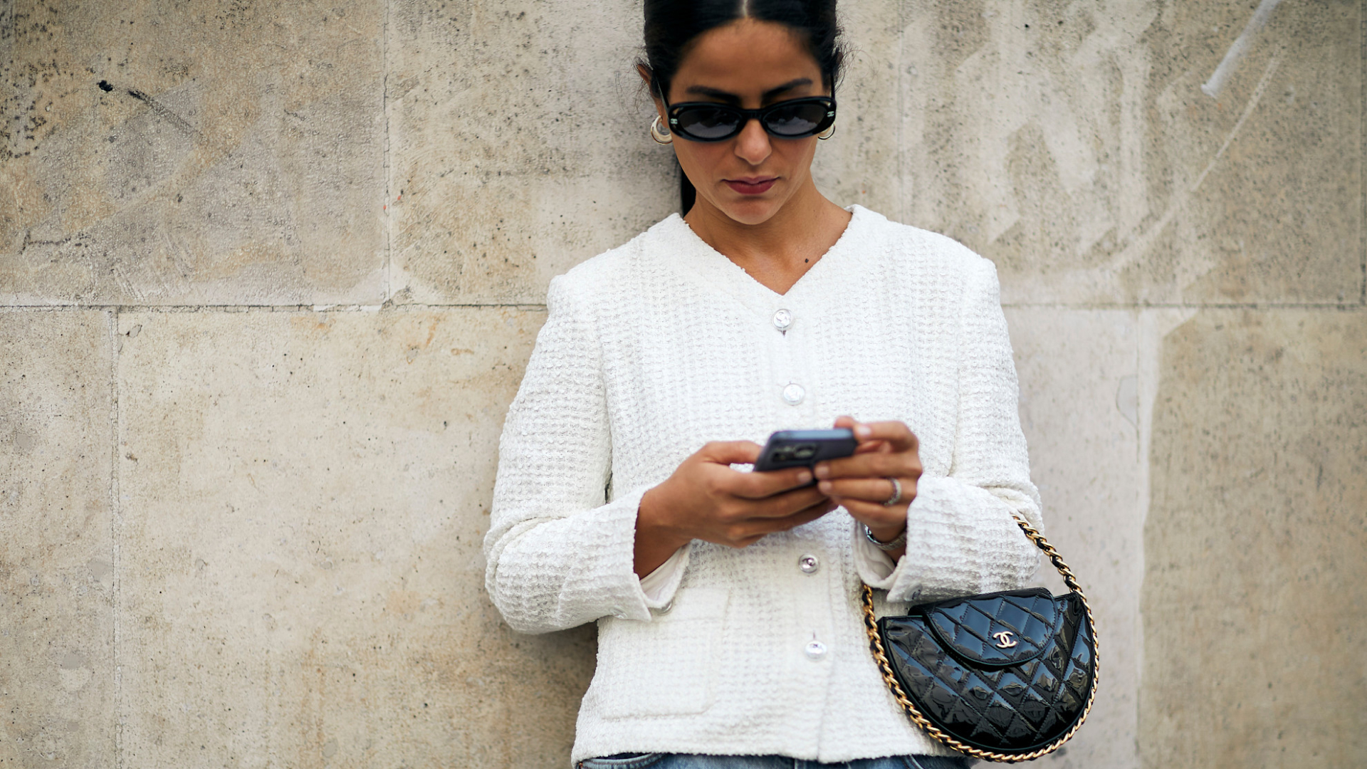 woman wearing white tweed jacket, sunglasses, and jeans looking at her phone and carrying a chanel bag