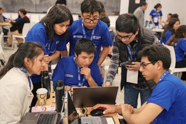 Six people look at a laptop computer in a classroom setting. Four wear blue t-shirts, one wears a white jacket, and one wears a plaid shirt with a black vest.