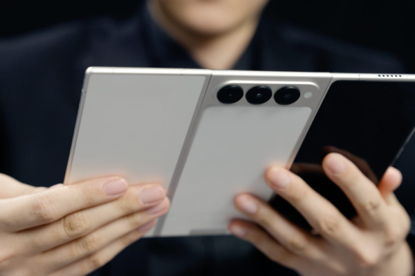 A person holds a tri-fold phone in its fully open state. We see the back, showing the three panels, three cameras, and a front screen.