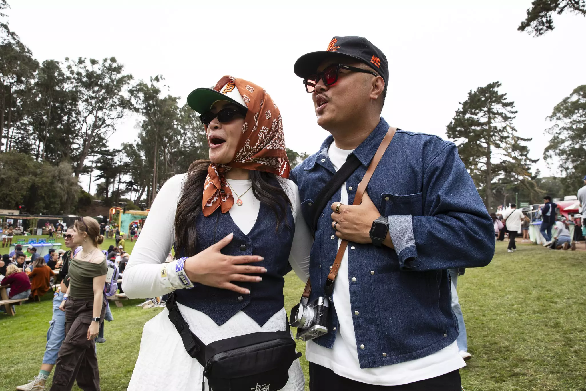 Two women wearing colorful, patterned clothing and sunglasses smile joyfully at an outdoor event, holding hands and embracing.