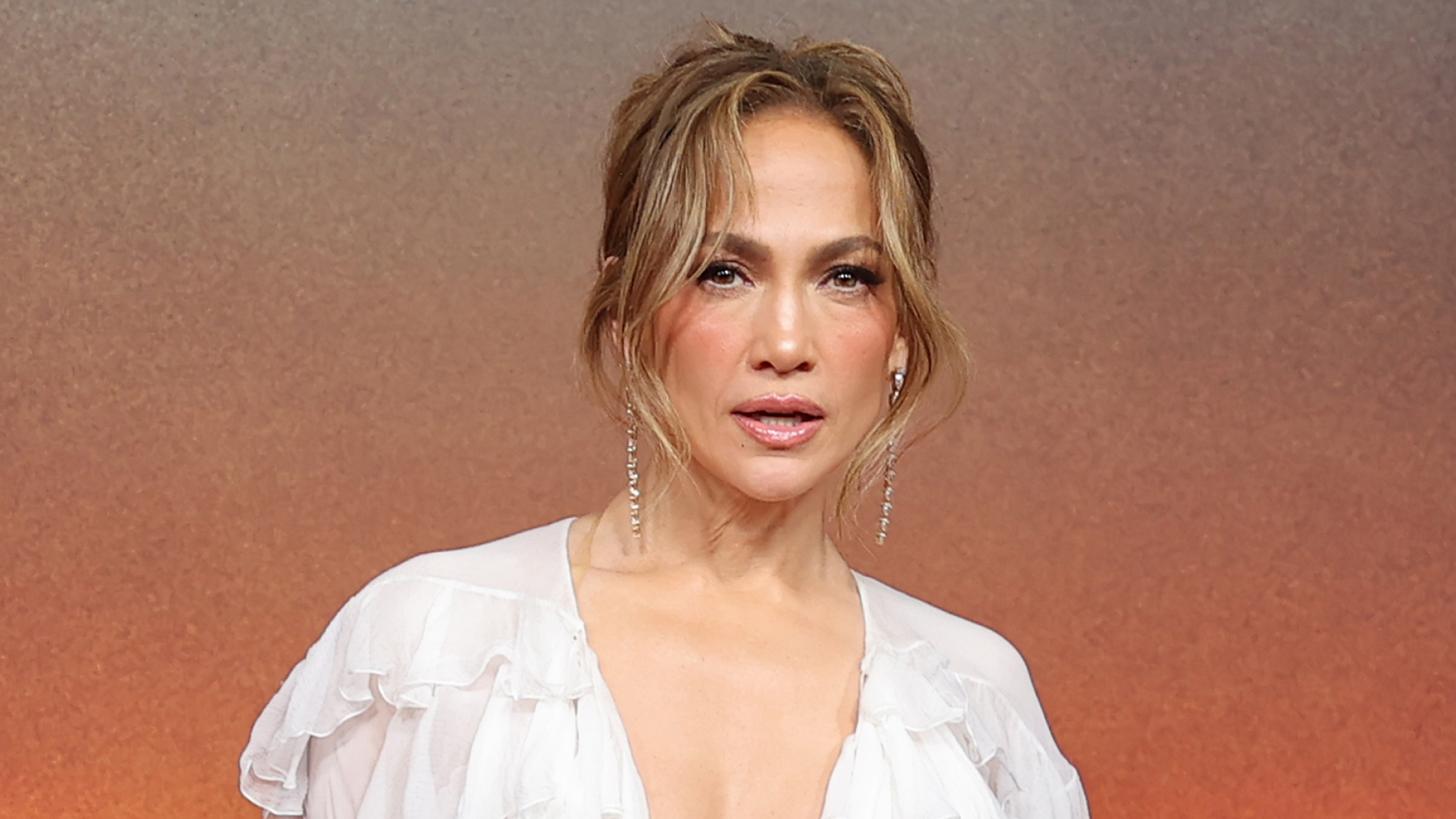 Jennifer Lopez wears a white tassled dress and stands in front of a sunset