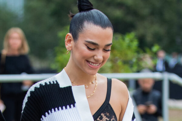 Dua Lipa has her hair in a high bun and wears an off-the-shoulder striped top with a lace bra underneath