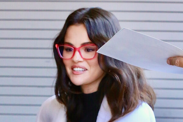Selena Gomez wearing heeled flip-flops, a black dress, a white coat, and red glasses