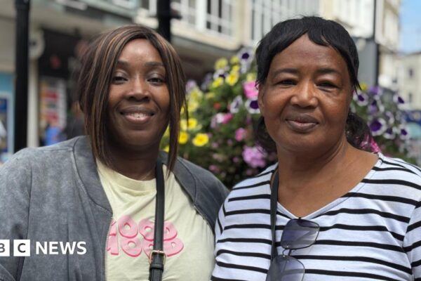 Two women - one in a blue and white striped long sleeved top with a handbag strap over her shoulder and sunglasses hanging off her collar, and the other in a yellow T-shirt and grey jacket with a crossbody bag strap visible, are standing on a high street and smiling at the camera. There are shops, floral displays and people blurred in the background.