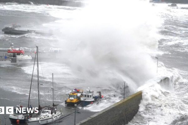Large white waves crashing into a harbour over small boats that are tied up