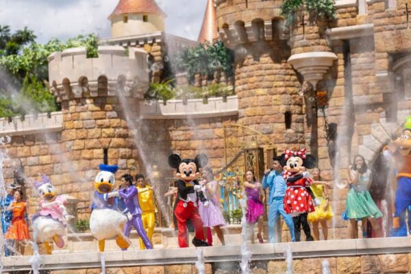 Colorful characters including Mickey, Minnie, Donald, Daisy, Goofy, and others dance in front of a castle with spouting fountains, entertaining guests on a sunny day at a theme park. The castle's detailed stonework is a prominent backdrop.