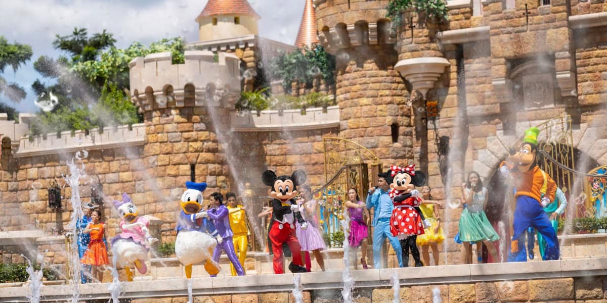 Colorful characters including Mickey, Minnie, Donald, Daisy, Goofy, and others dance in front of a castle with spouting fountains, entertaining guests on a sunny day at a theme park. The castle's detailed stonework is a prominent backdrop.