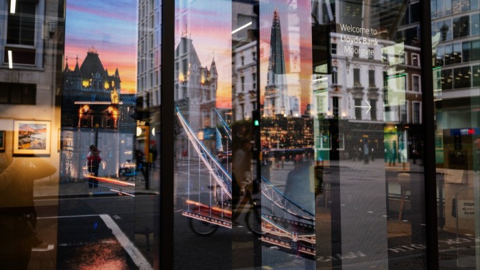 Reflections of city buildings and street scenes blend with an illuminated display of London landmarks on the glass frontage of a Lloyds Bank branch.