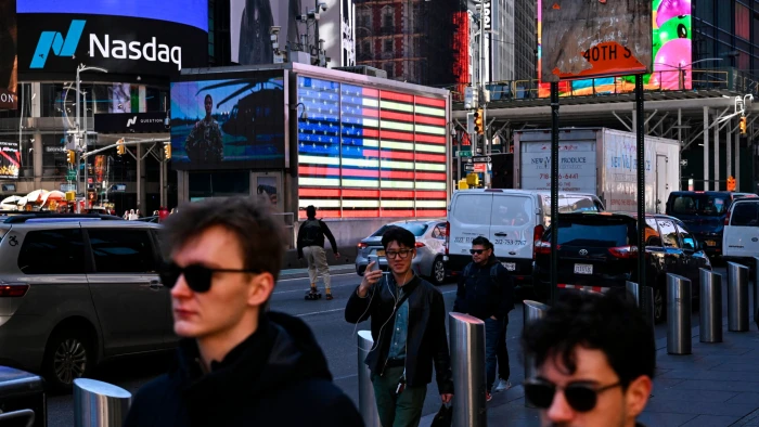 Pedestrians pass by the Nasdaq MarketSite and a large American flag display in Times Square amid busy city traffic