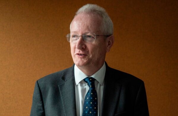 Niall Mackenzie, Acas chief executive, wearing a suit and patterned tie, stands against a plain brown background.