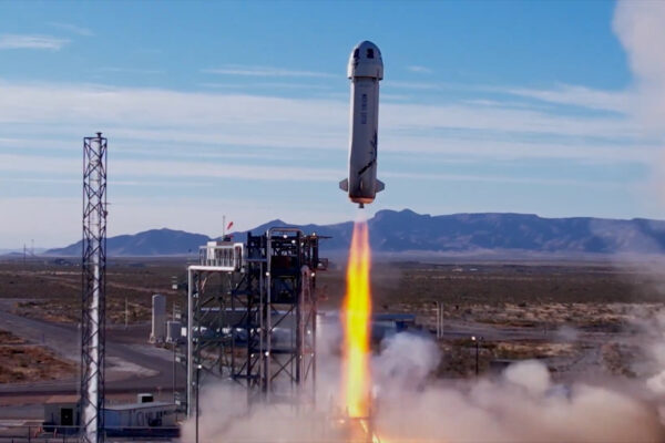 A screenshot from a YouTube video showing Blue Origin's New Shepard rocket blasting away from its launch pad against a blue sky