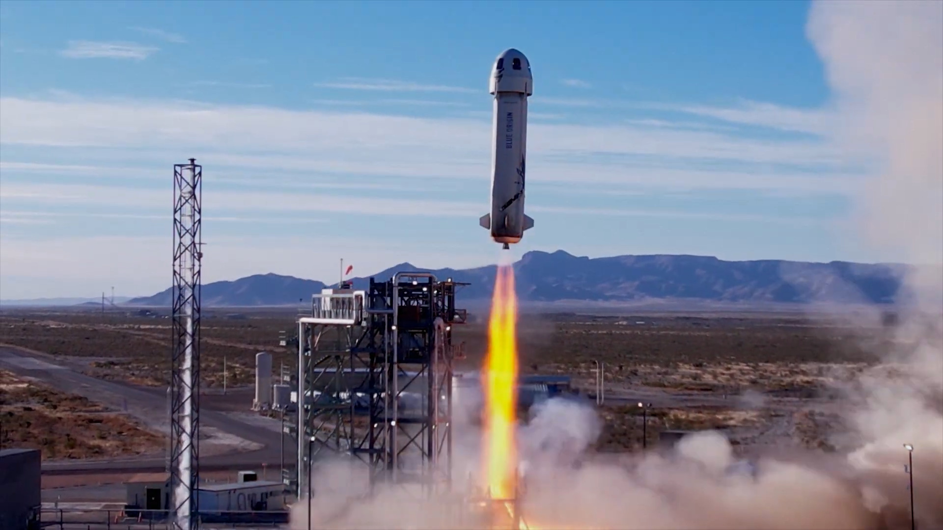 A screenshot from a YouTube video showing Blue Origin's New Shepard rocket blasting away from its launch pad against a blue sky